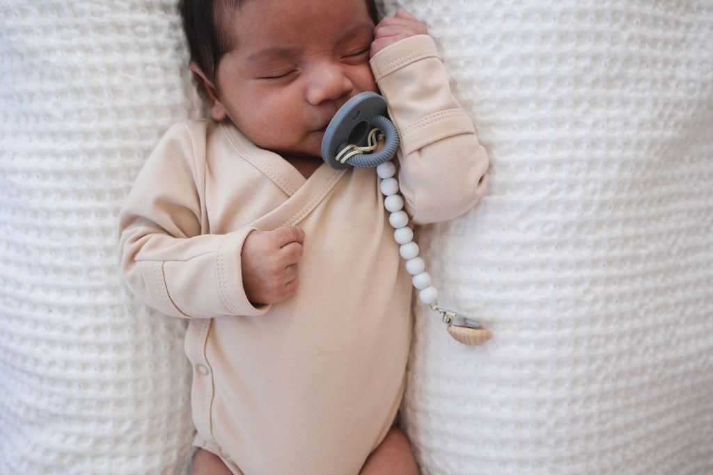 Newborn baby in a beige onesie holding a pacifier with a white textured blanket in the background