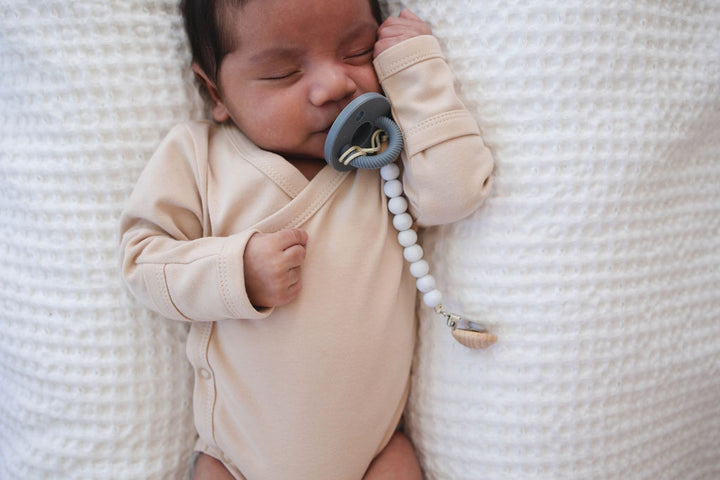 Newborn baby in a beige onesie holding a pacifier with a white textured blanket in the background