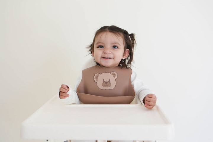 Child wearing a brown bib with a bear design, sitting in a high chair against a plain background